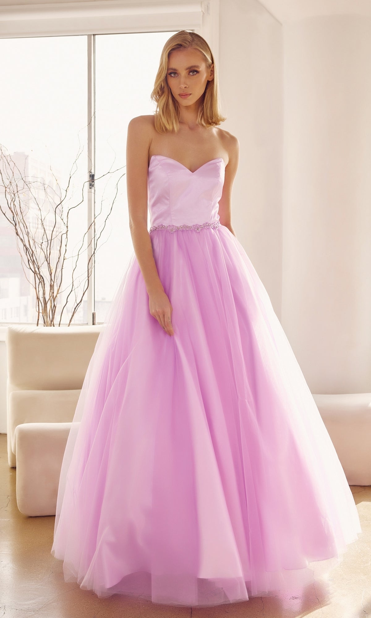 A woman stands indoors in the Strapless Sweetheart Long Prom Ball Gown 265, featuring a fitted bodice, corset-style back, and full light pink tulle skirt. Natural light streams through a window behind her.